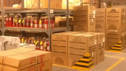 Warehouse storage area with stacked cardboard boxes and shelves filled with fire extinguishers under bright lighting
