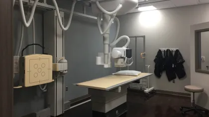 Modern X-ray room with a digital radiography machine, examination table, and protective lead aprons on the wall.