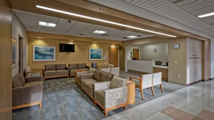 Modern medical waiting room with beige chairs, wood accents, blue carpet, and a reception desk with clocks.