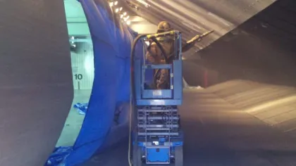 Worker in lift cleaning or coating large metallic aircraft wing inside hangar under artificial lighting.