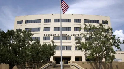 Front view of Keesler Medical Center building with American flag and surrounding trees under blue sky.