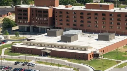 A large brick institutional building with multiple stories and a white flat roof, surrounded by parking and greenery.