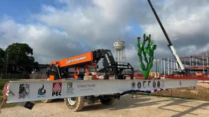 Construction site with a large white steel beam, logos, an orange lift, and a cactus balloon under cloudy sky.