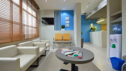 Modern waiting room with white chairs, yellow seats, magazines on table, large window blinds, and water dispenser.