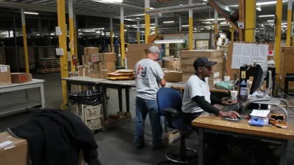 Workers packing boxes and using a computer in a busy warehouse with shelves and packages around.