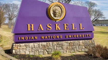 Purple and gold Haskell Indian Nations University entrance sign with Native American chief emblem on stone base.