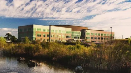 Modern office building with large windows near a pond and grassy foreground under a cloudy blue sky.
