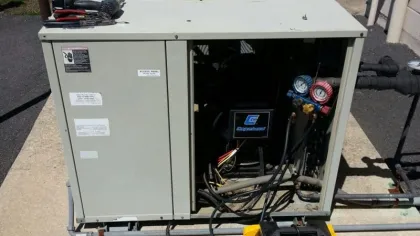 Commercial HVAC unit with open panel showing gauges and tools set on top, outdoors on concrete pad.