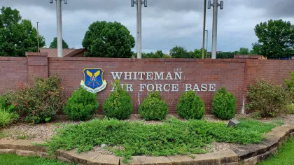 Brick entrance sign for Whiteman Air Force Base with emblem, greenery, and overcast sky in background.