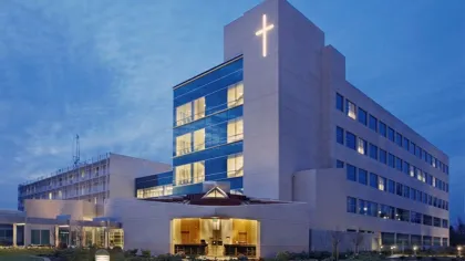 Modern hospital building with illuminated cross at dusk, featuring large windows and landscaped surroundings.