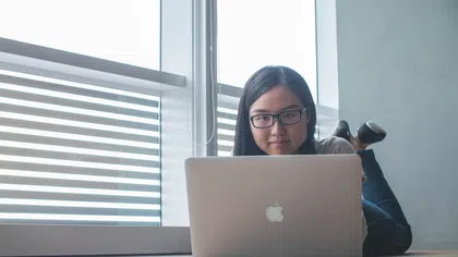 Young woman wearing glasses lying on the floor working on a MacBook laptop near window with blinds.
