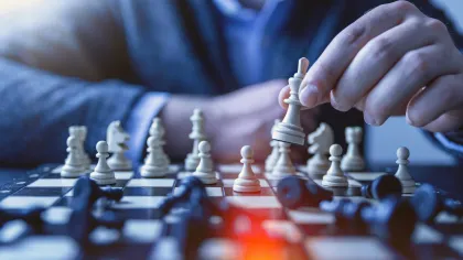 Person in blue sweater moves white king piece in a strategic chess game on a checkered board.