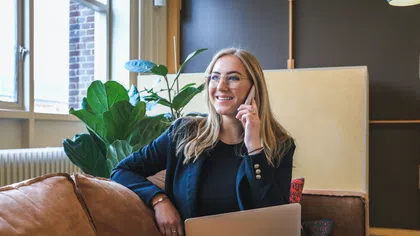 Young professional woman smiling and talking on phone while sitting on couch with laptop in modern office.