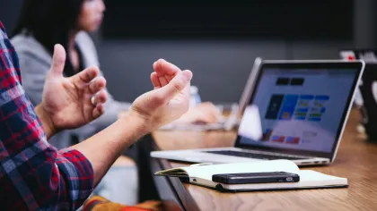 Person gesturing during a meeting with a laptop, notebook, and smartphone on the wooden table.