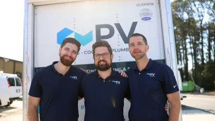 Three men wearing matching navy blue polo shirts with PV Heating Cooling Plumbing logo stand in front of a service truck.