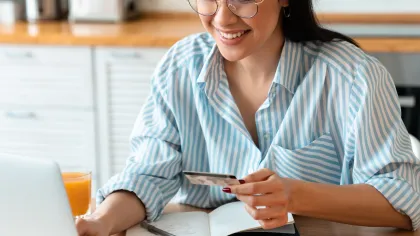 Smiling woman using laptop for online shopping while holding a credit card in a bright kitchen.