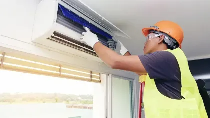 Worker wearing safety gear repairing or installing a split air conditioning unit indoors near a window.
