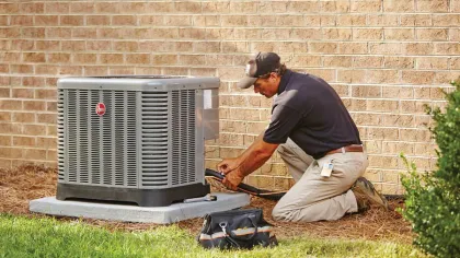 Technician kneeling by an outdoor HVAC unit installing or repairing it against a brick wall on grass.