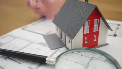 Model house, magnifying glass, and piggy bank on architectural blueprints for home planning and savings.