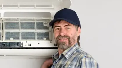 Technician in plaid shirt and cap repairing or servicing a wall-mounted air conditioning unit indoors.