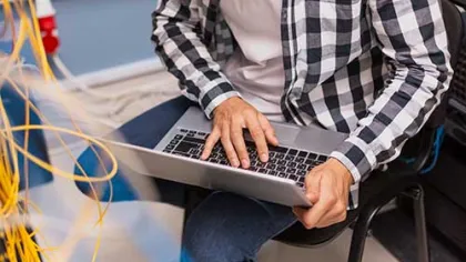 Person using a laptop while sitting near network cables in a tech workspace or server room.