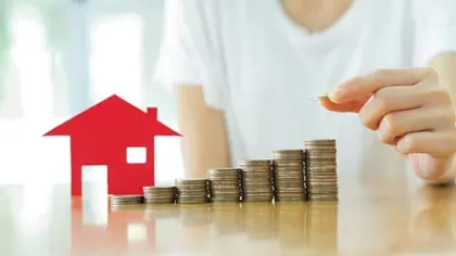 Person stacking coins in ascending order next to a red house model symbolizing real estate investment and savings growth.