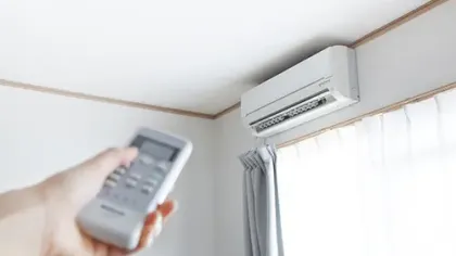 Hand holding remote control pointing at white wall-mounted air conditioner in bright room.