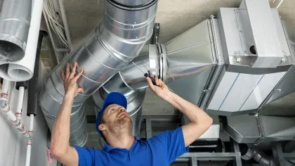 Technician in blue uniform inspecting and repairing metal HVAC ductwork with a screwdriver in a commercial building.