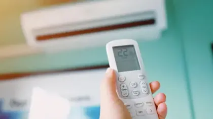 Hand holding remote control set to 22°C in front of a wall-mounted air conditioning unit.