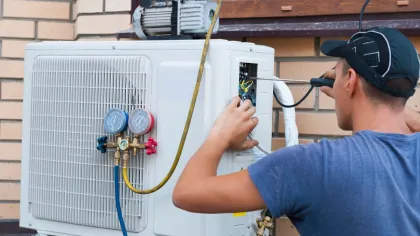 Technician repairing and inspecting an outdoor air conditioning unit with gauges and tools near a brick wall.
