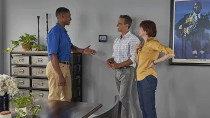 Technician greeting a smiling couple inside a modern home near a smart thermostat on the wall.