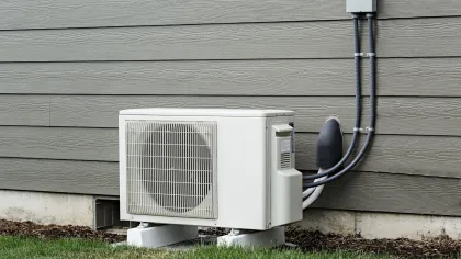 Outdoor air conditioning unit installed on concrete blocks beside a gray house with grass around the base.