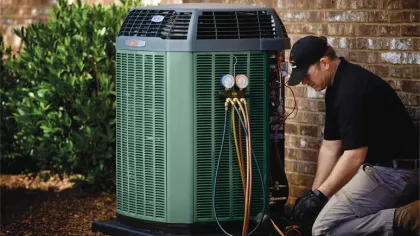 Technician servicing a green outdoor air conditioning unit attached to a brick wall with gauges and cables.