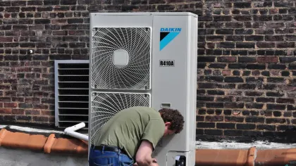 Technician repairing or installing Daikin R410A air conditioning unit on rooftop with brick wall background.