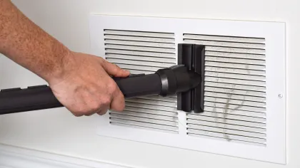 Hand using vacuum cleaner attachment to clean dust from a white air vent grille on a wall.