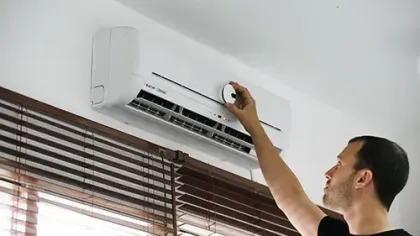 Man adjusting the thermostat of a wall-mounted air conditioner inside a room with wooden blinds.