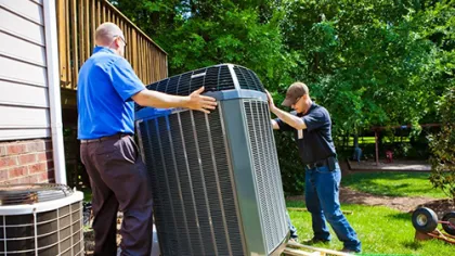 Two HVAC technicians installing a large outdoor air conditioning unit next to a house on a sunny day.