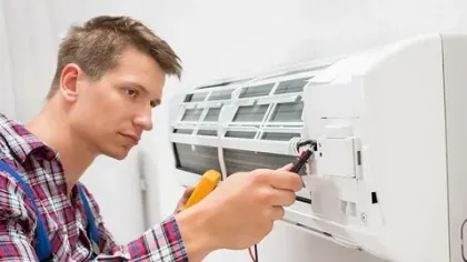 Technician in plaid shirt repairing and testing a wall-mounted air conditioner with a multimeter tool.