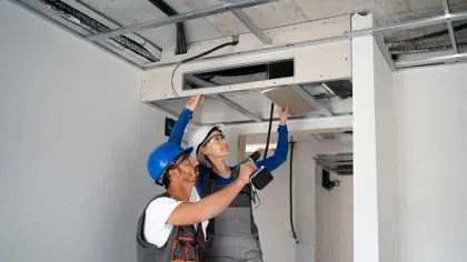Two construction workers installing ceiling panels inside a building under renovation with safety helmets and tools.