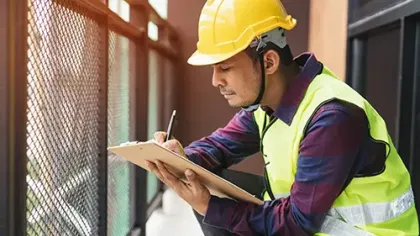 Construction worker in yellow helmet and vest taking notes on clipboard outdoors near fence in daylight.