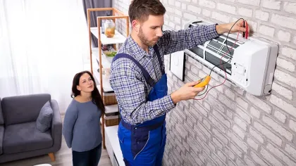 Technician in blue overalls repairing wall-mounted air conditioner while woman watches in living room.