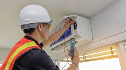 Technician in safety gear using multimeter to test a wall-mounted air conditioner indoors near a window.