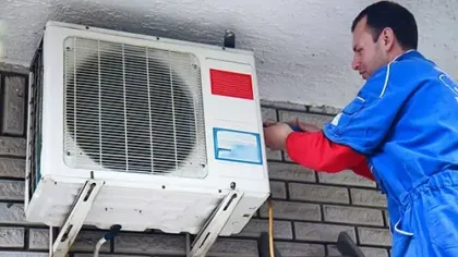 Technician installing or repairing an outdoor air conditioning unit mounted on a brick wall.