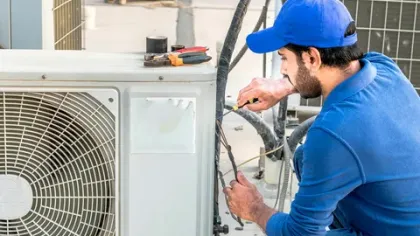 Technician in blue uniform repairing outdoor air conditioning unit with tools on top during daytime.