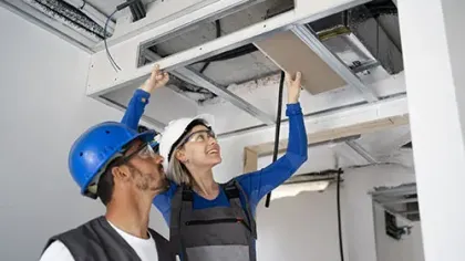 Two construction workers wearing helmets inspecting and installing building framework in an unfinished ceiling.