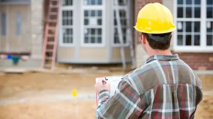 Construction worker in hard hat inspecting house building site with clipboard near brick wall and windows.