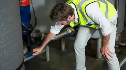 Worker in safety vest adjusting valve on industrial piping system in a mechanical room.
