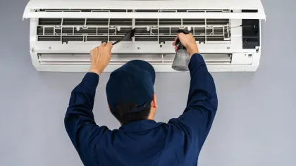 Technician cleaning and maintaining a wall-mounted air conditioner using a brush and spray bottle
