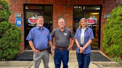 Three plumbing professionals in uniform stand in front of Rooter Plus office doors with company logos visible.