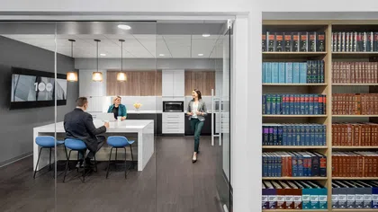 Modern office kitchen with glass walls, three people conversing, blue chairs, and a bookshelf filled with legal books.